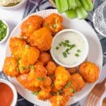 A large white plate is full of red-orange air fryer buffalo cauliflower. They are plated alongside a small white bowl of ranch dipping sauce and covered with finely chopped green chives. Nearby, a bowl of green celery sticks and a small bowl of buffalo sauce adorn the image.
