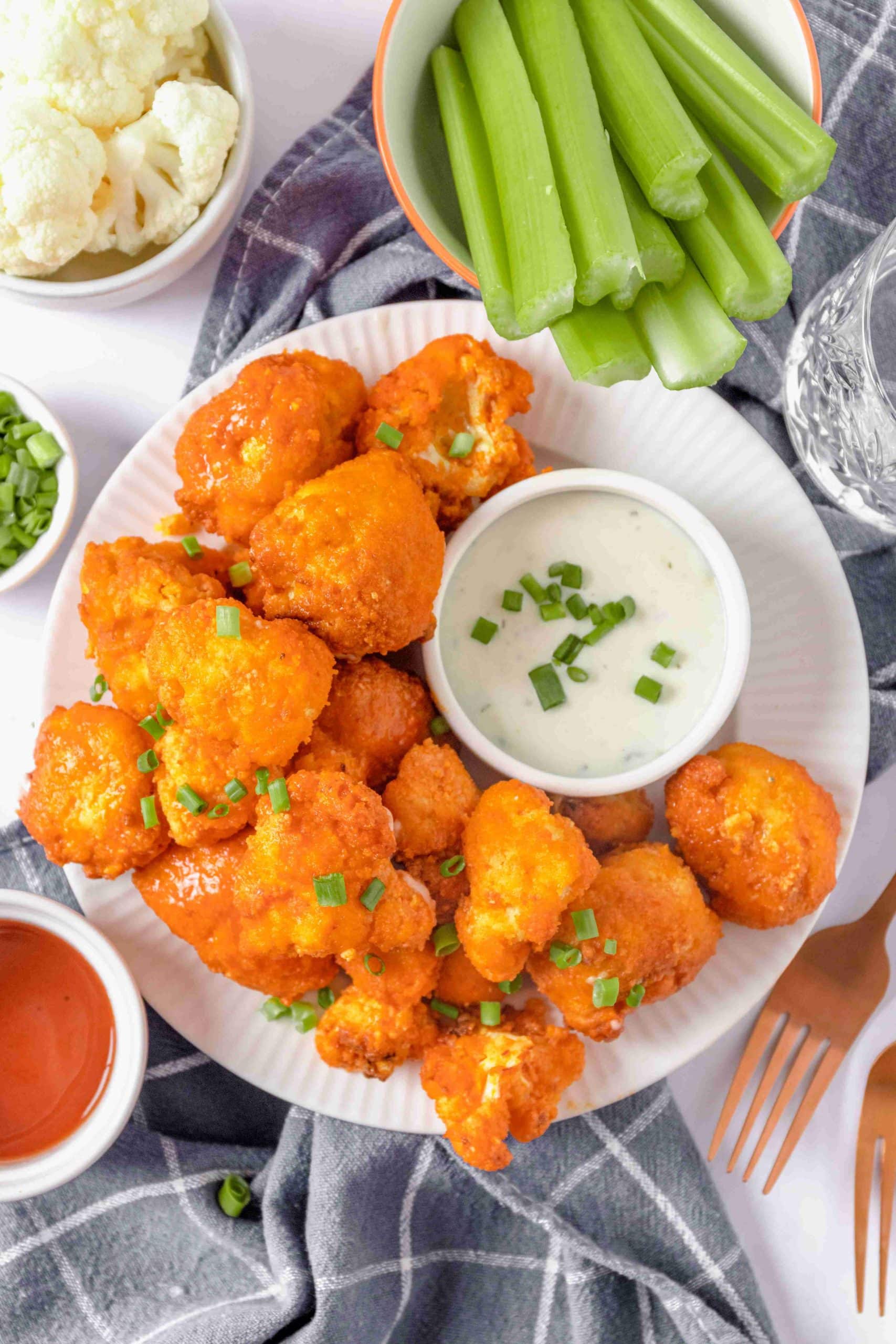 A large white plate is full of red-orange air fryer buffalo cauliflower. They are plated alongside a small white bowl of ranch dipping sauce and covered with finely chopped green chives. Nearby, a bowl of green celery sticks and a small bowl of buffalo sauce adorn the image.