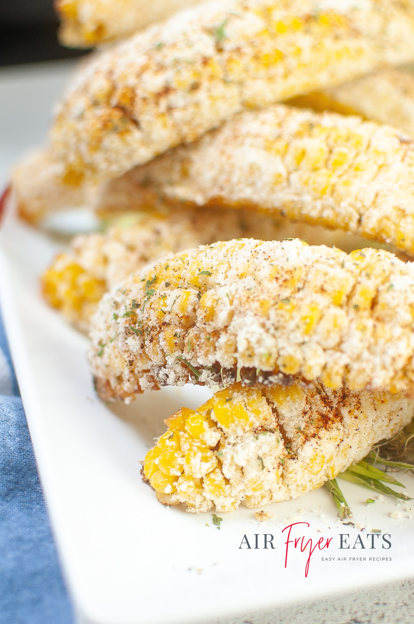 Golden-yellow Mexican street corn made in the air fryer is covered with white Parmesan cheese, red spices, and green flakes. The corn pieces are piled on top of each other on a white plate.