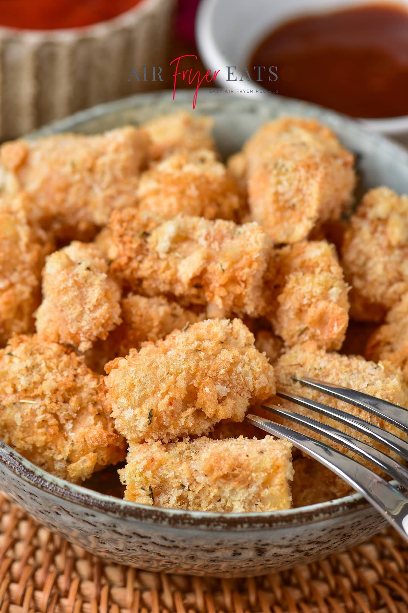 Close-up shot of homemade air fryer tater tots piled in a ceramic bowl. The tater tots are crispy and covered in golden-brown breadcrumbs. A silver fork lies on the side of the bowl.