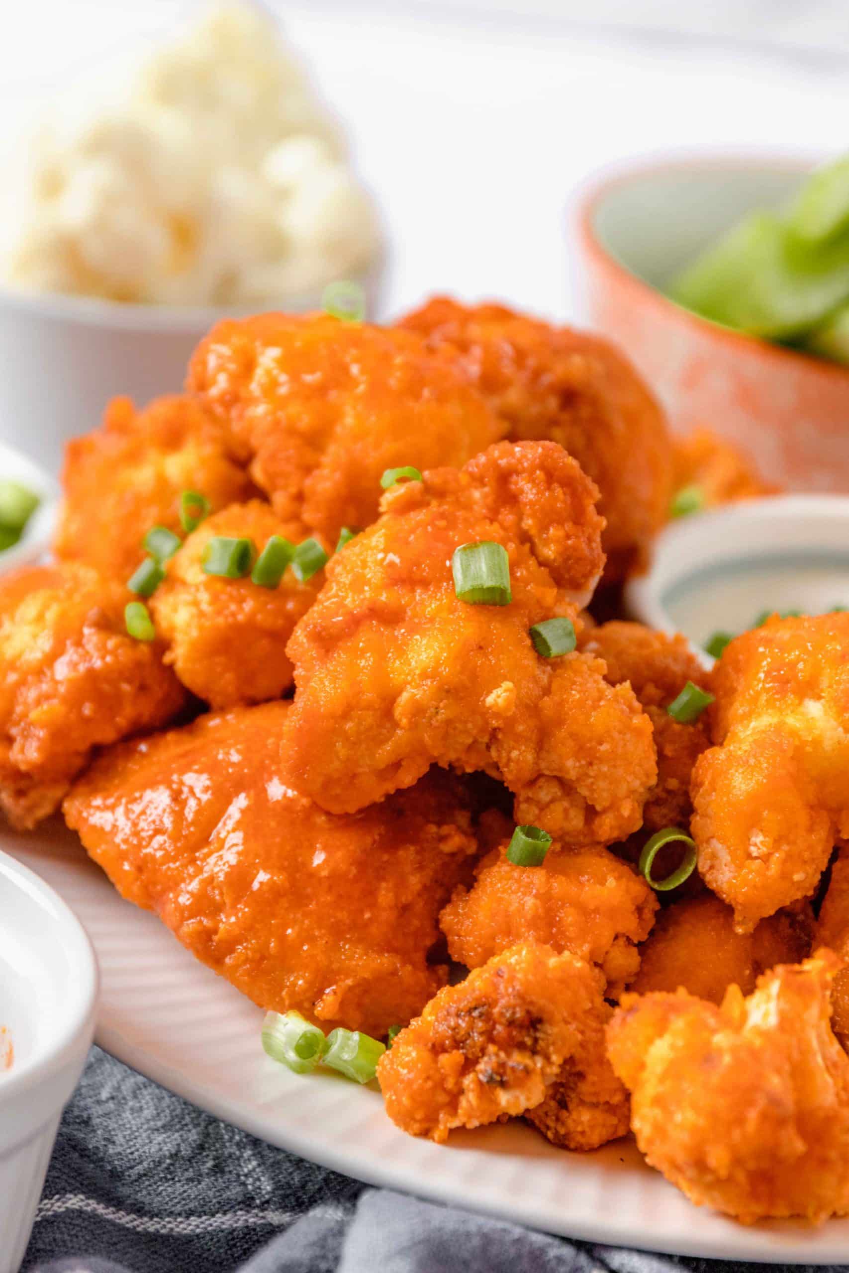 A pile of red-orange buffalo cauliflower is covered with finely-chopped green chives. There are raw cauliflower and celery sticks in the background.