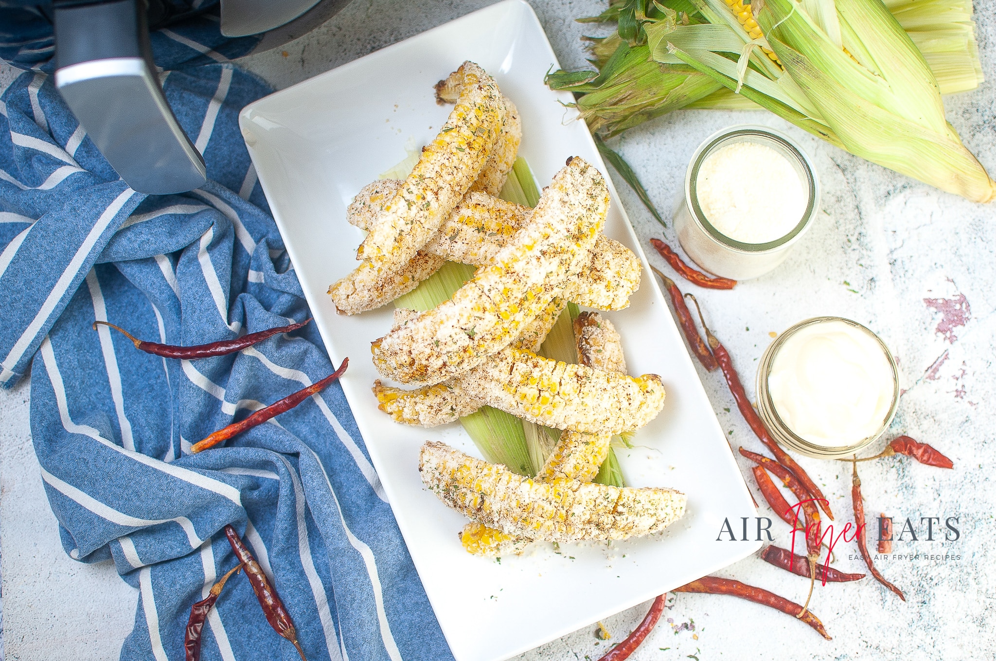 Elote corn covered in parmesan cheese and spices is piled on a white plate. The raw corn, parmesan cheese, and mayonnaise ingredients surround the plate. There are red chilies surrounding the plate on the kitchen counter, and there is an air fryer above the plate.