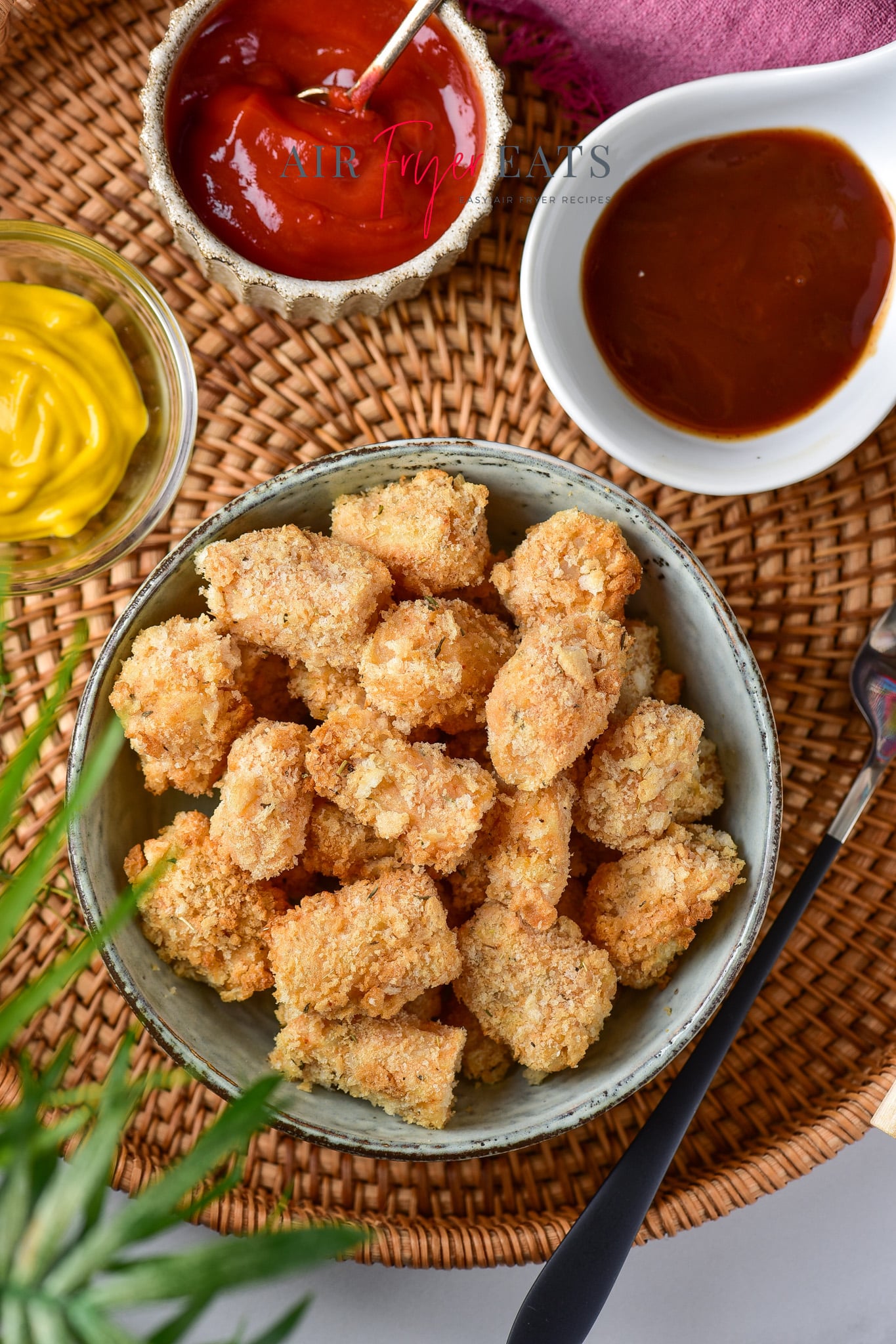 Crispy homemade tater tots air fryer are piled in a ceramic bowl. Nearby, ketchup, barbecue sauce, and mustard are seen in smaller bowls.