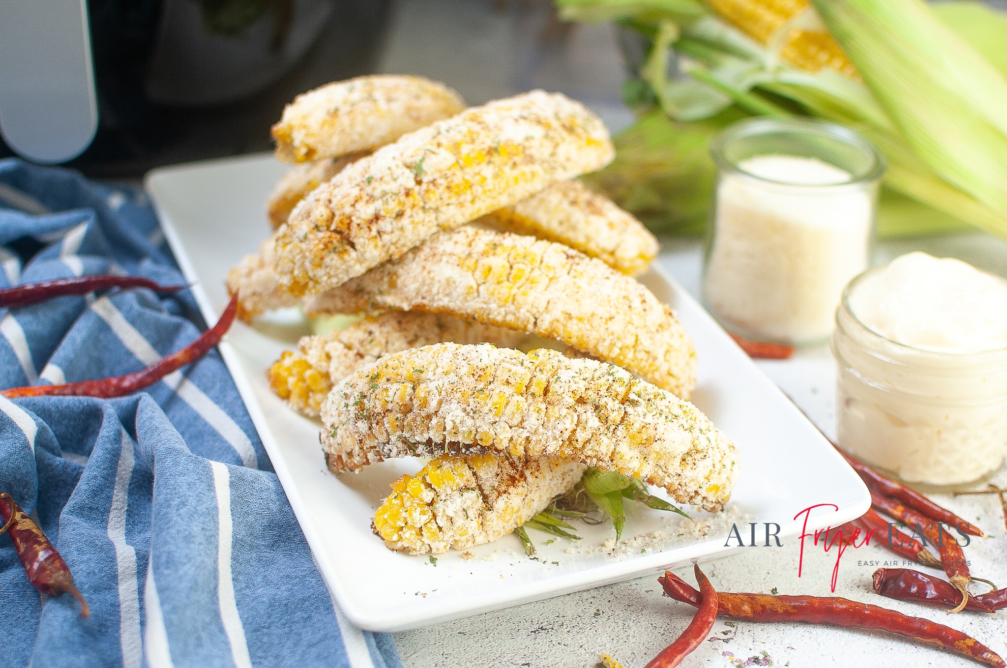 A plate full of golden-yellow Mexican street corn made in the air fryer makes up the center of the image. The corn is covered with mayonnaise, parmesan cheese, and spices. Ingredients like corn, parmesan cheese, mayonnaise, and red chilis surround the plate. An air fryer can be seen in the background.