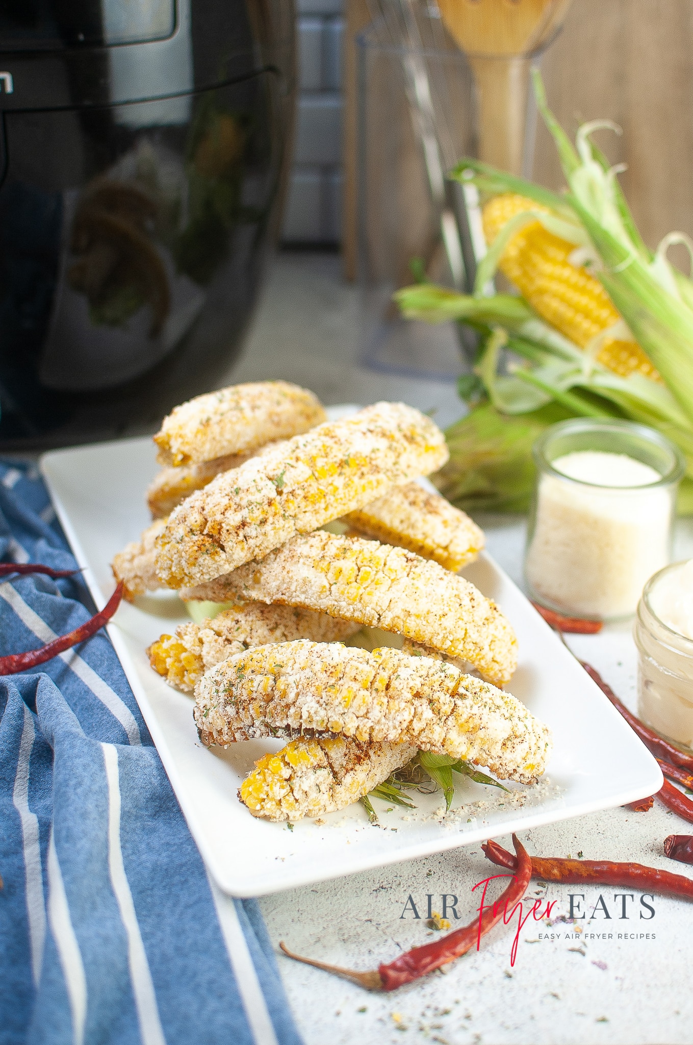Vertical image of a plate with golden-yellow Mexican street corn made in the air fryer stacked on top of each other. Corn, chilies, cheese, and mayonnaise surround the plate on the kitchen counter.