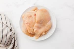 Thick, raw chicken breasts are on a white plate on a marble kitchen countertop. There is a black and white striped towel on the counter as well.