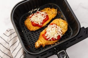 Two breaded parmesan chicken cutlets lie in an air fryer basket. There is red marinara sauce and freshly grated mozarella cheese on top of each chicken breast.