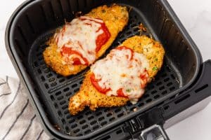 Two cooked, breaded parmesan chicken cutlets lie in an air fryer basket. They are crispy, golden-brown, and topped with red marinara sauce and melted mozarella cheese.