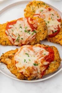 Three chicken parmesan cutlets topped with marinara sauce and mozarella cheese are placed on a white plate on a white kitchen surface.