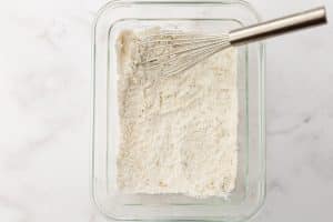 A shallow glass dish contains a flour mixture. A silver whisk rests inside the dish.