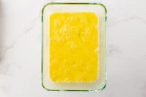 A glass dish is filled with beaten eggs. The egg mixture is yellow and has multiple bubbles of various sizes. The dish is on a white marble countertop.