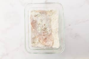 A flour-coated chicken breast lies in a glass dish inside a flour mixture.