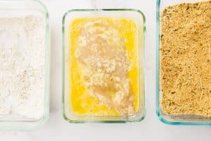 A flour-coated chicken breast lies in a glass dish with an egg coating. On the left, there is a glass dish filled with a flour mixture. On the right, a glass dish filled with a breadcrumbs and parmesan cheese mixture.