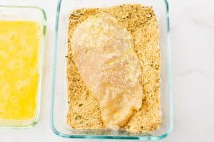 A flour-and-egg-coated chicken breast lies in a glass dish with a parmesan cheese mixture. There is a glass dish with an egg mixture on the left of the dish.