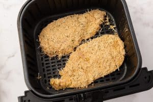 Two raw, breaded chicken breasts lie in an air fryer basket.