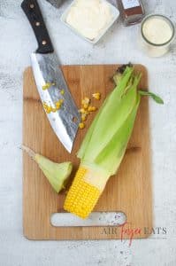 A corn cob is halfway covered with corn husk on a cutting board. A large knife has cut off the end of the corn husk, and there are corn kernels spread on the cutting board.