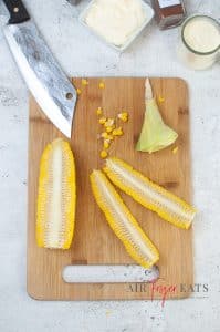 Two slices of long corn pieces are laid on a cutting board. The remaining corn is also on the cutting board. A large knife is resting halfway on the cutting board.