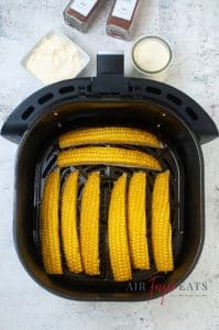 Eight pieces of yellow corn are placed in an air fryer basket. The corn is cut lengthwise into quarter pieces. The corn is bright yellow.