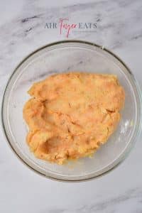A dough-like mass of potato mixed with spices and cornstarch sits in a glass bowl on a marble kitchen counter.