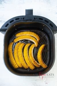 Eight pieces of cooked corn are in an air fryer basket. The corn is bright yellow and has barts that are cooked brown from the heat of the air fryer.
