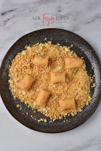 Small, uncooked tater tot pieces lay on a black plate full of breadcrumbs on a marble kitchen countertop.