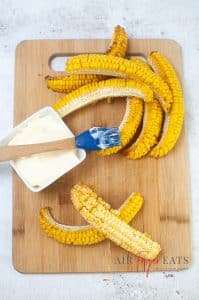 Pieces of cooked corn are laid on a cutting board. There is a cooking brush laid on top of a dish of mayonnaise that is used to brush the corn pieces with mayonnaise.