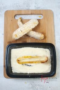 A cooked corn piece is laid in a dish with grated Parmesan cheese. There are two other corn pieces that are covered in Parmesan cheese laid on a cutting board.