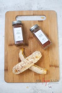 Pieces of cooked corn covered in mayonnaise, Parmesan cheese, chili powder and cayenne pepper are placed on a cutting board. Two spice bottles filled with chili powder and cayenne pepper are laid on the cutting board.