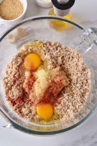 A glass mixing bowl is full of a grey turkey mixture. Atop the ground turkey are two raw eggs, an onion mixture, and spices.