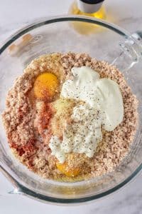 A glass mixing bowl is full of a grey turkey mixture. Atop the ground turkey are two raw eggs, greek yogurt, an onion mixture, and spices.