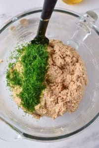 A glass mixing bowl is full of a grey turkey mixture. Freshly chopped green dill is added to the mixture. There is a black mixing spatula inside the bowl.