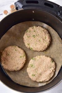 Three uncooked turkey patties are placed on parchment paper in a circular air fryer basket.