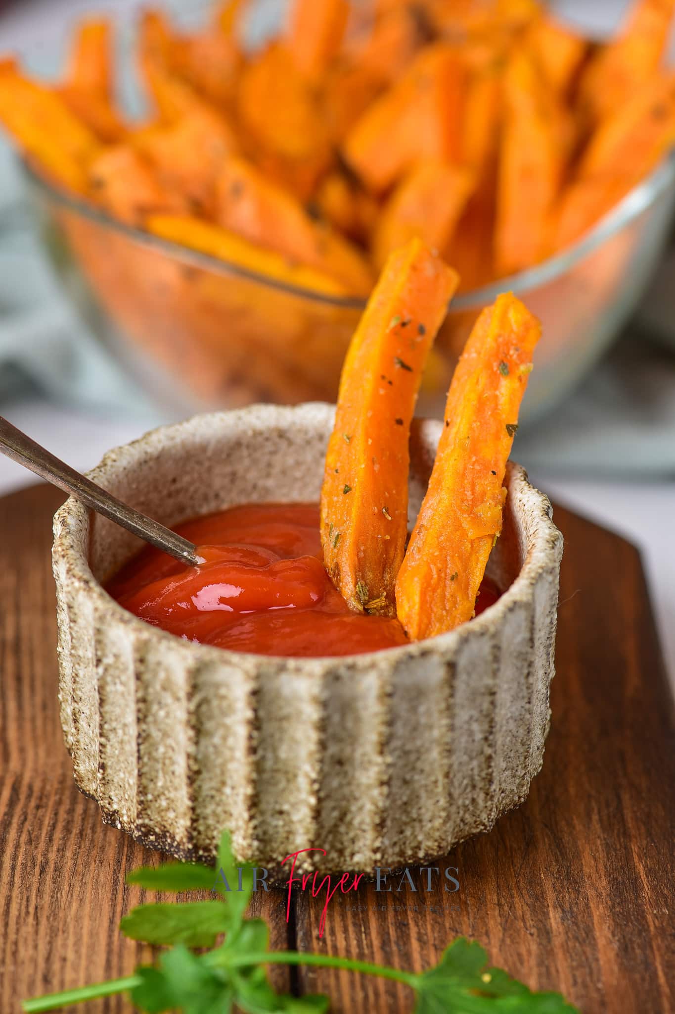 Two air fryer sweet potato fries are propped up in a small dish filled with ketchup. A large bowl of sweet potato fries can be seen in the background.