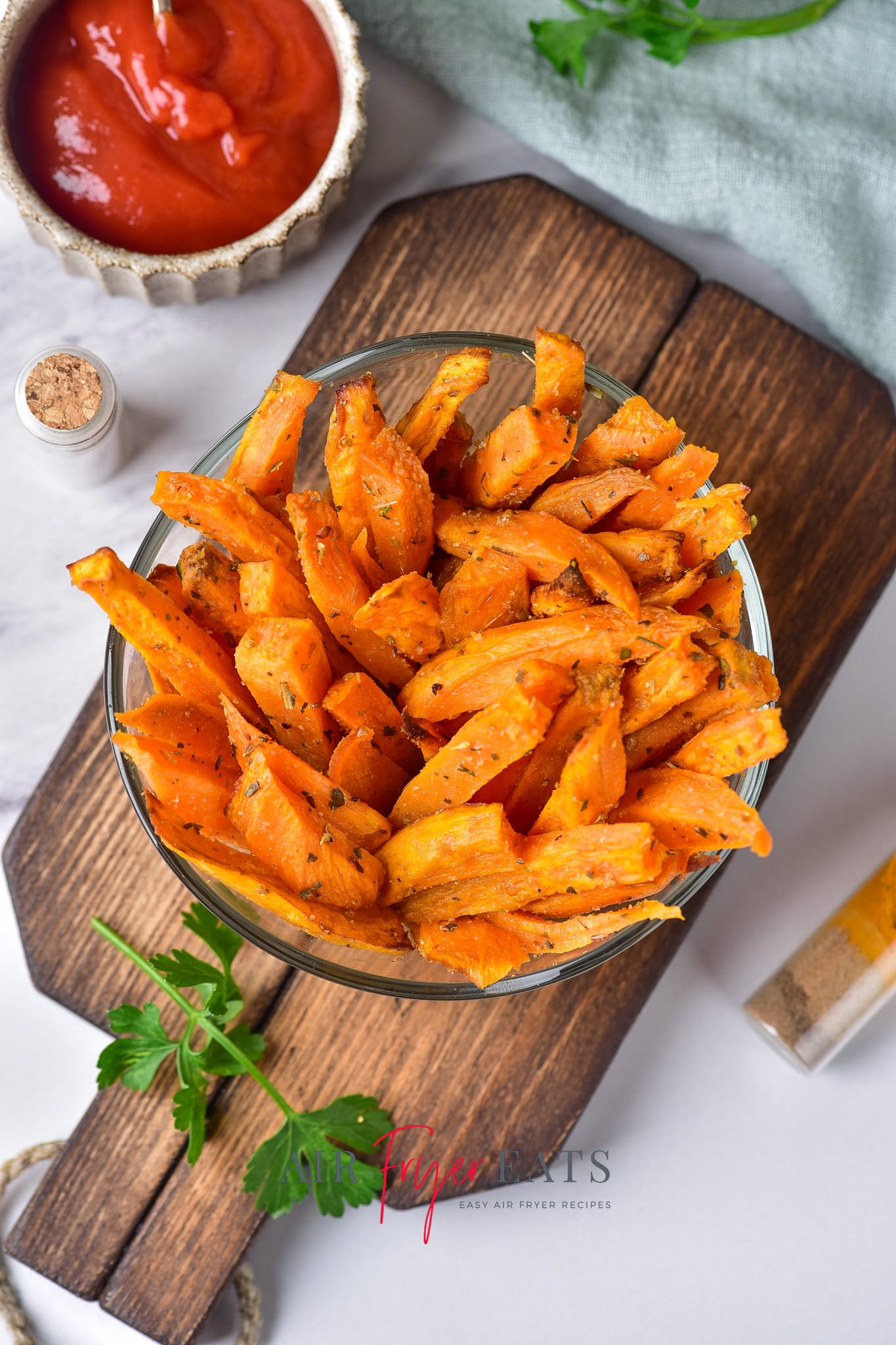 A large bowl is full of crispy air fryer sweet potato fries. They are bright orange and covered in spices. It is placed on a wooden board and ketchup can be seen in the distance.