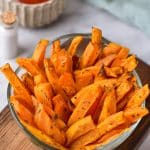 A large bowl is full of crispy air fryer sweet potato fries. They are bright orange and covered in spices. There is a bowl of ketchup in the background for dipping the fries.