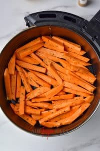 Slices of uncooked sweet potatoe fries are covered in spices and olive oil and sit in the bottom of a circular air fryer basket.