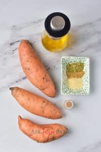 Three orange raw sweet potatos are placed on a kitchen counter alongside a small dish of spices and a flask of olive oil.