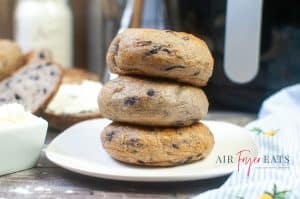 Three air fryer blueberry bagels are stacked on a white plate. A large, black air fryer can be seen in the background of the photo. The bagels are brown and speckled with purple blueberries.