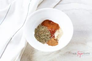 A small white bowl is filled with various spices: oregano, smoked paprika, garlic salt, and cinnamon.