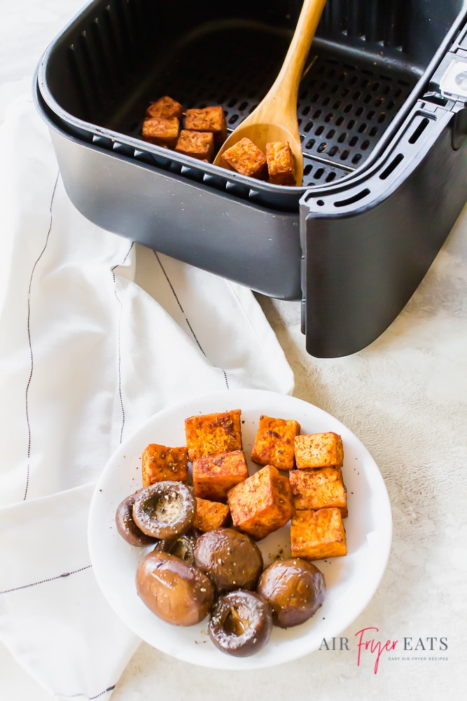 Air fryer smoky tofu is served with a wooden spoon from a rectangular air fryer basket. The crispy tofu is orange in color with golden spots. Next to the air fryer basket, there is a small white plate filled with crispy tofu cubes and balsamic mushrooms.