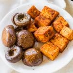 A close-up shot of a white plate filled with medium-sized crispy tofu cubes that are a rich orange color and brown balsamic mushrooms covered with salt and pepper.