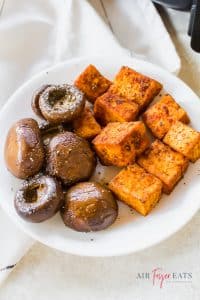 A close-up shot of a white plate filled with medium-sized crispy tofu cubes that are a rich orange color and brown balsamic mushrooms covered with salt and pepper.