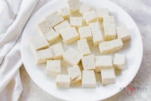 A white plate is full of freshly cubed tofu. The tofu bites are medium-sized and have small holes in them. They are placed next to a white kitchen towel.
