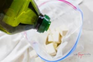 A close-up shot of a green bottle of olive oil being poured into a plastic bag filled with cubed tofu.