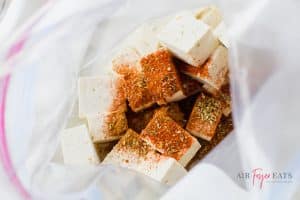 A close-up shot of a bag of firm tofu cubes covered with mixed spices. The spices are red and green, smoked paprika and oregano can be seen.