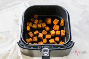 Cooked tofu bites are in a square-shaped air fryer basket. They are orange and brown and crispy on the edges.