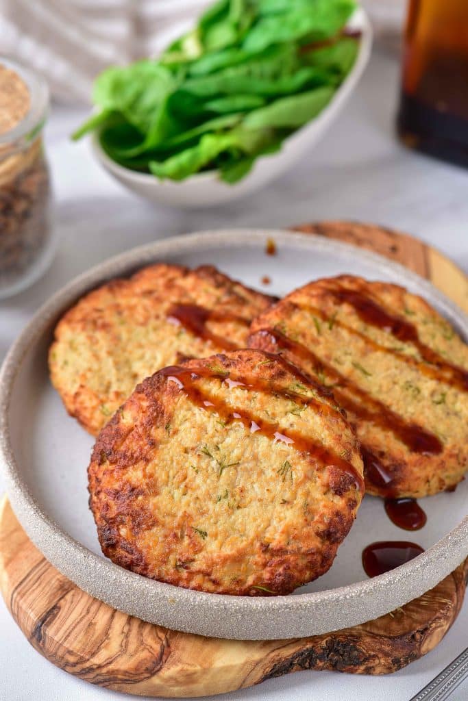 Three golden-brown air fryer turkey patties are placed on a gray plate. They are covered with a little bit of barbecue sauce. In the background, there is fresh green lettuce.