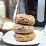 Three air fryer blueberry bagels are stacked on a white plate. A large, black air fryer can be seen in the background of the photo. The bagels are brown and speckled with purple blueberries.