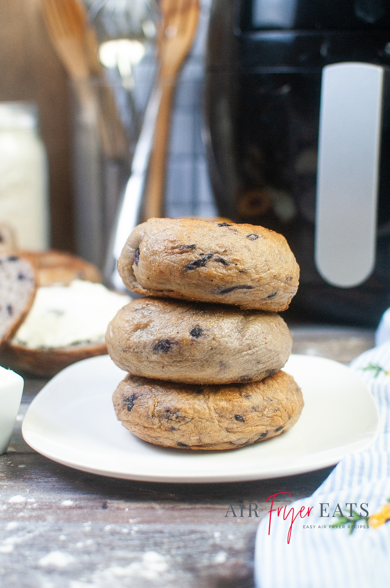 Three air fryer blueberry bagels are stacked on a white plate. A large, black air fryer can be seen in the background of the photo. The bagels are brown and speckled with purple blueberries.