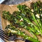 Air fryer broccolini are placed on a brown wax paper on a grey plate. The broccolini are crispy and have dark green florets and are covered with parmesan and garlic. A black fork is on the plate.